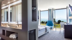 A modern hotel room with a large mirror, a bathtub, a bed with a view, a desk, and a chair. - Starling Hotel Lausanne (Saint-Sulpice)
