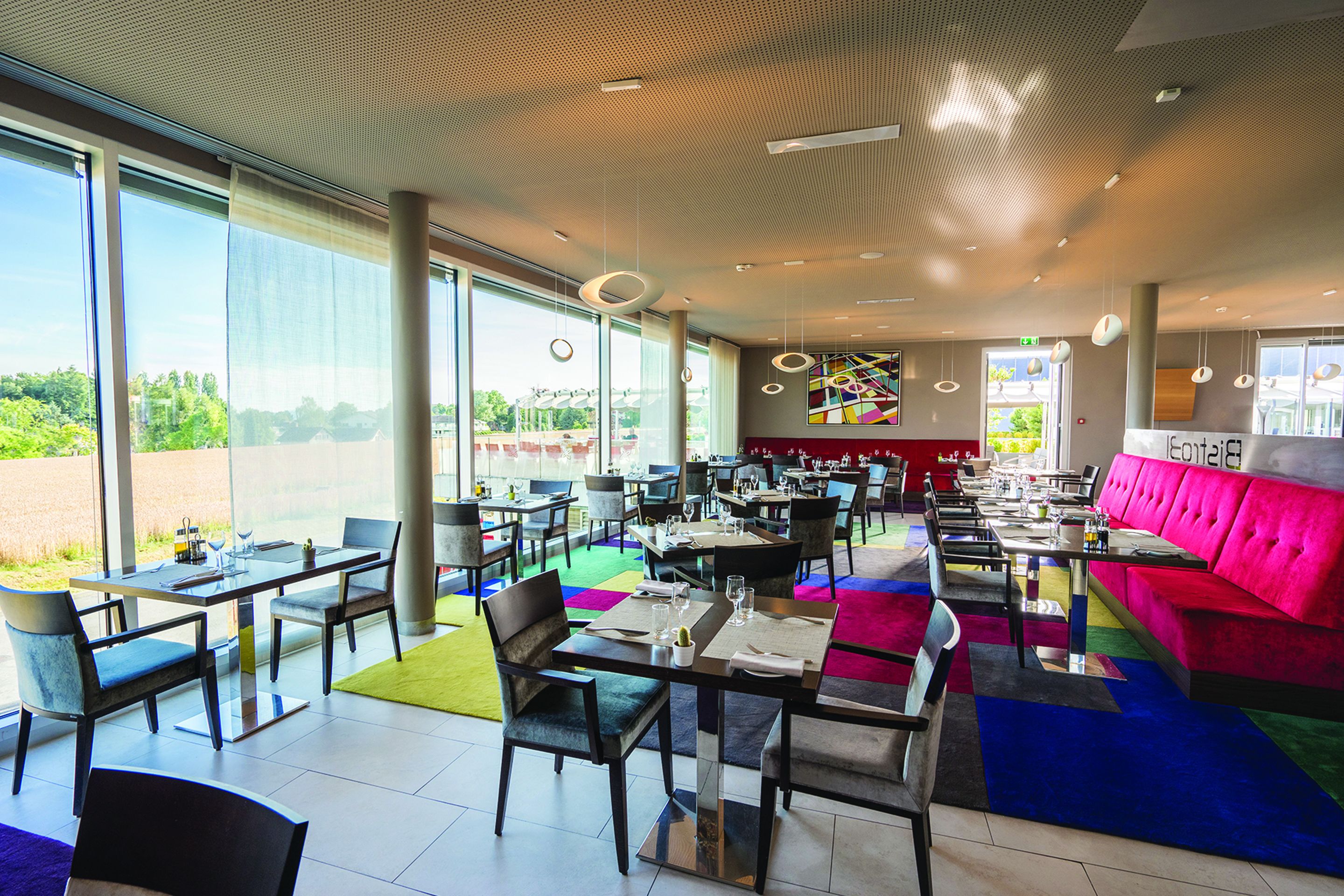 A modern restaurant with large windows, a colorful carpet, and a red sofa.