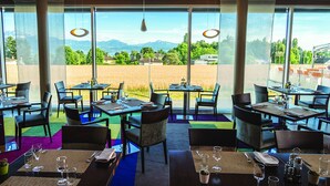 A restaurant with tables set for dining, large windows offering a view of a field and mountains, and hanging pendant lights. - Starling Hotel Lausanne (Saint-Sulpice)