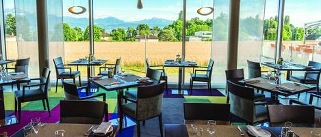 A restaurant with tables set for dining, large windows offering a view of a field and mountains, and hanging pendant lights.