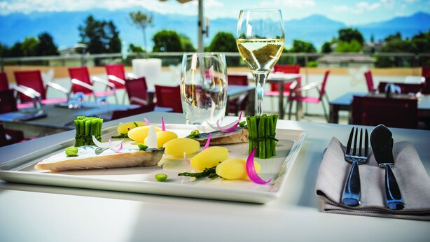 A dining table with a white plate of food, a glass of white wine, and a napkin with cutlery. - Starling Hotel Lausanne (Saint-Sulpice)