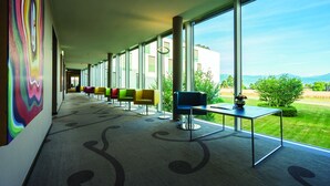 A modern hotel lobby with a long corridor, colorful chairs, a table with a vase, and large windows offering a view of greenery and mountains. - Starling Hotel Lausanne (Saint-Sulpice)