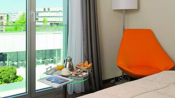 Une chambre d’hôtel avec un lit, une table où sont disposés des aliments pour le petit-déjeuner, une chaise orange vif et une vue sur les bâtiments à travers de grandes fenêtres.