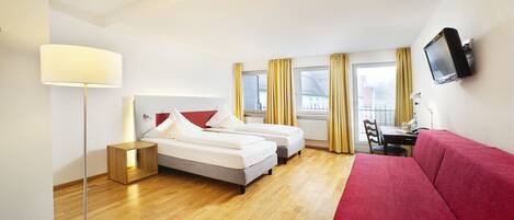 A hotel room with a bed, a red sofa, a desk with a chair, a TV mounted on the wall, and a window with curtains.