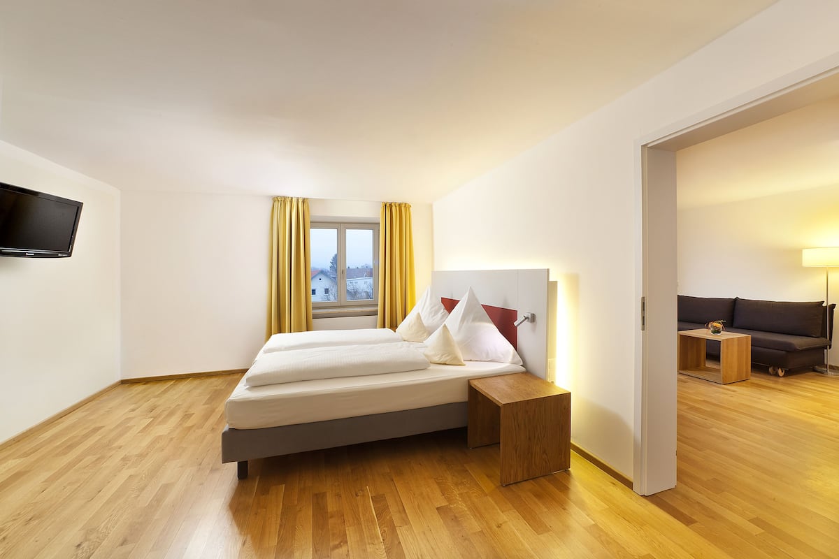 Une chambre d’hôtel moderne équipée d’un lit, d’une télévision, d’une fenêtre avec des rideaux, d’un plancher en bois et d’un canapé dans la pièce adjacente.
