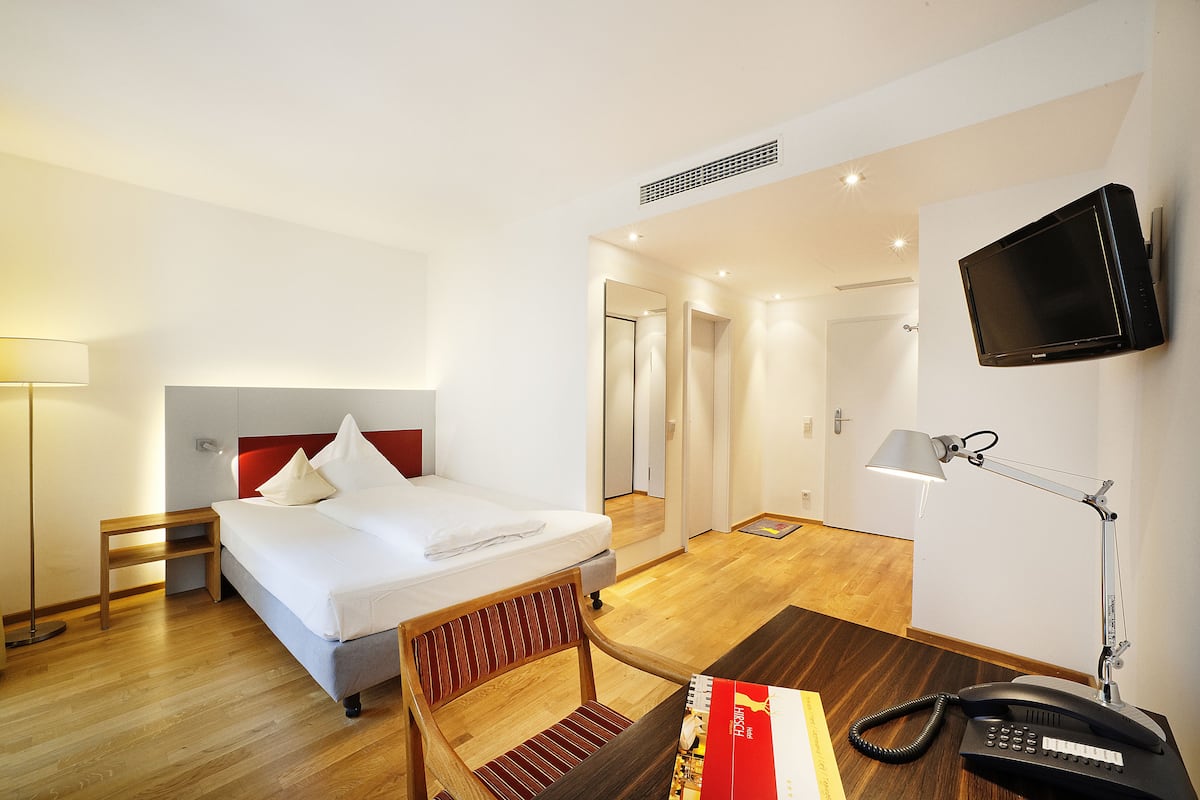 Une chambre d’hôtel comprenant un lit, un bureau avec un téléphone, une chaise, une lampe de bureau et un téléviseur fixé au mur.