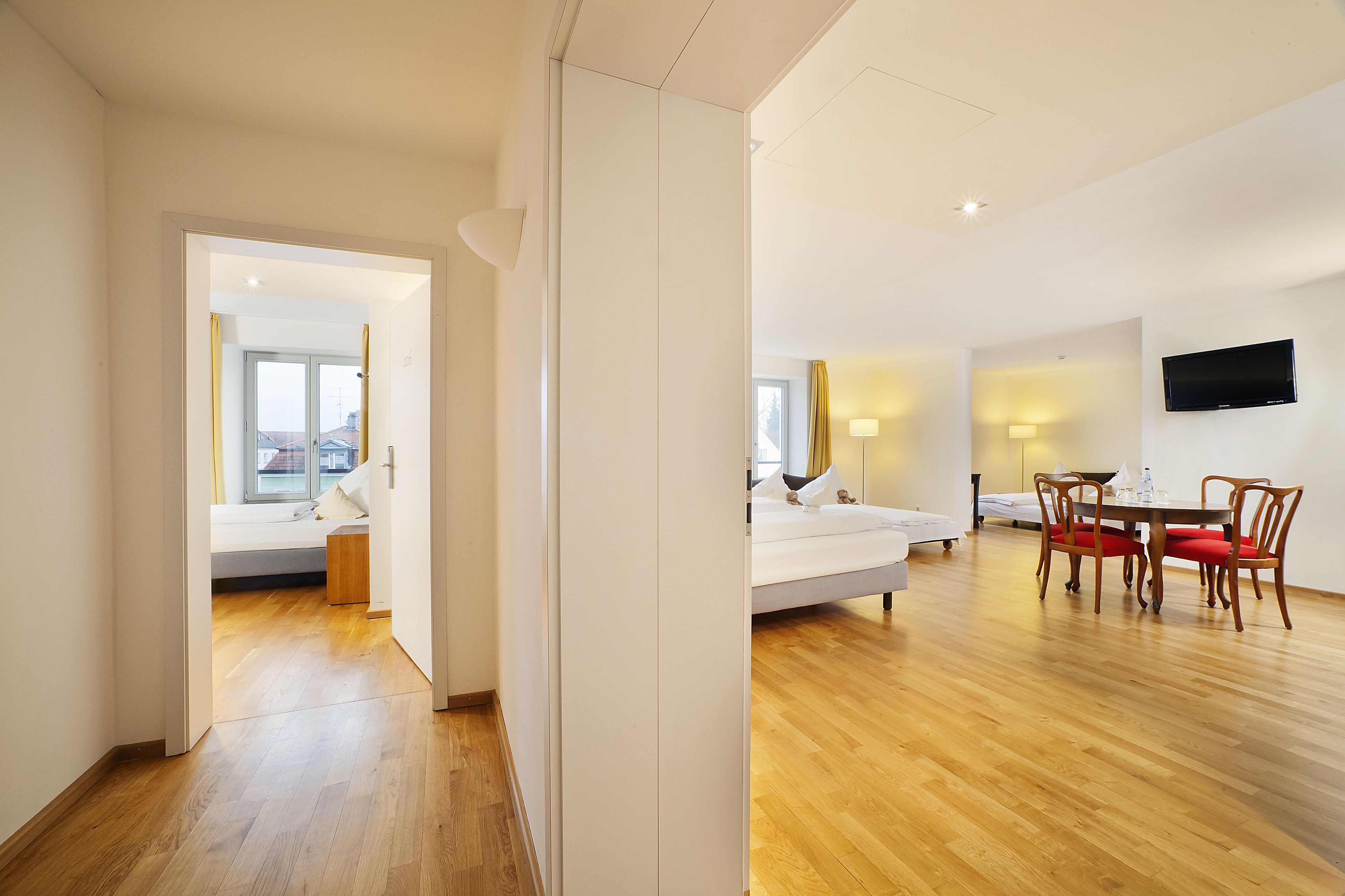 Ein modernes Hotelzimmer mit Holzboden, einem Bett, einer Essecke mit Tisch und Stühlen sowie einem an der Wand befestigten Fernseher.