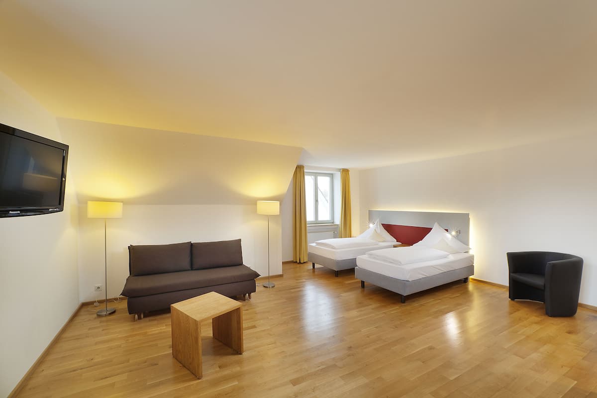 Une chambre d’hôtel moderne avec un plancher en bois, un canapé, deux lits simples, une petite table, une télévision et une fenêtre avec des rideaux.