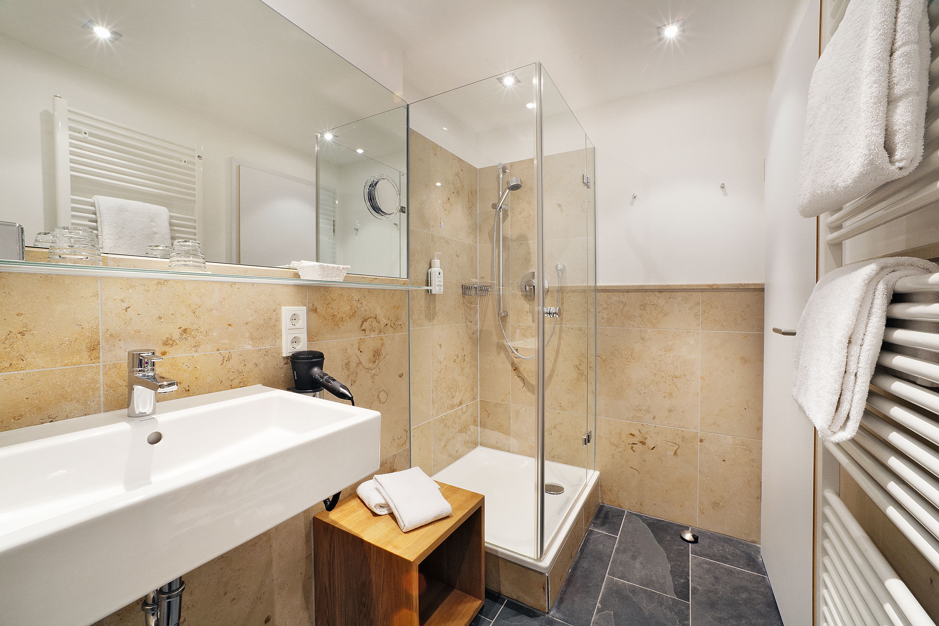 Een moderne badkamer met een glazen douchecabine, een witte wastafel en een houten krukje.