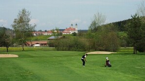 Ein Golfplatz mit einem Bunker, eine Person in Schwarz, die bereit ist, einen Golfball zu schlagen, und eine weitere Person in Weiß, die auf dem Rasen sitzt.