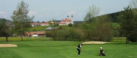 Ein Golfplatz mit einem Bunker, eine Person in Schwarz, die bereit ist, einen Golfball zu schlagen, und eine weitere Person in Weiß, die auf dem Rasen sitzt.