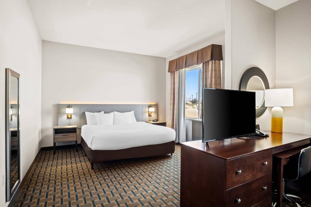 Ein Hotelzimmer mit einem großen Bett, einem Schreibtisch, einem Flachbildfernseher, einer Lampe und einem fenster mit Vorhängen.