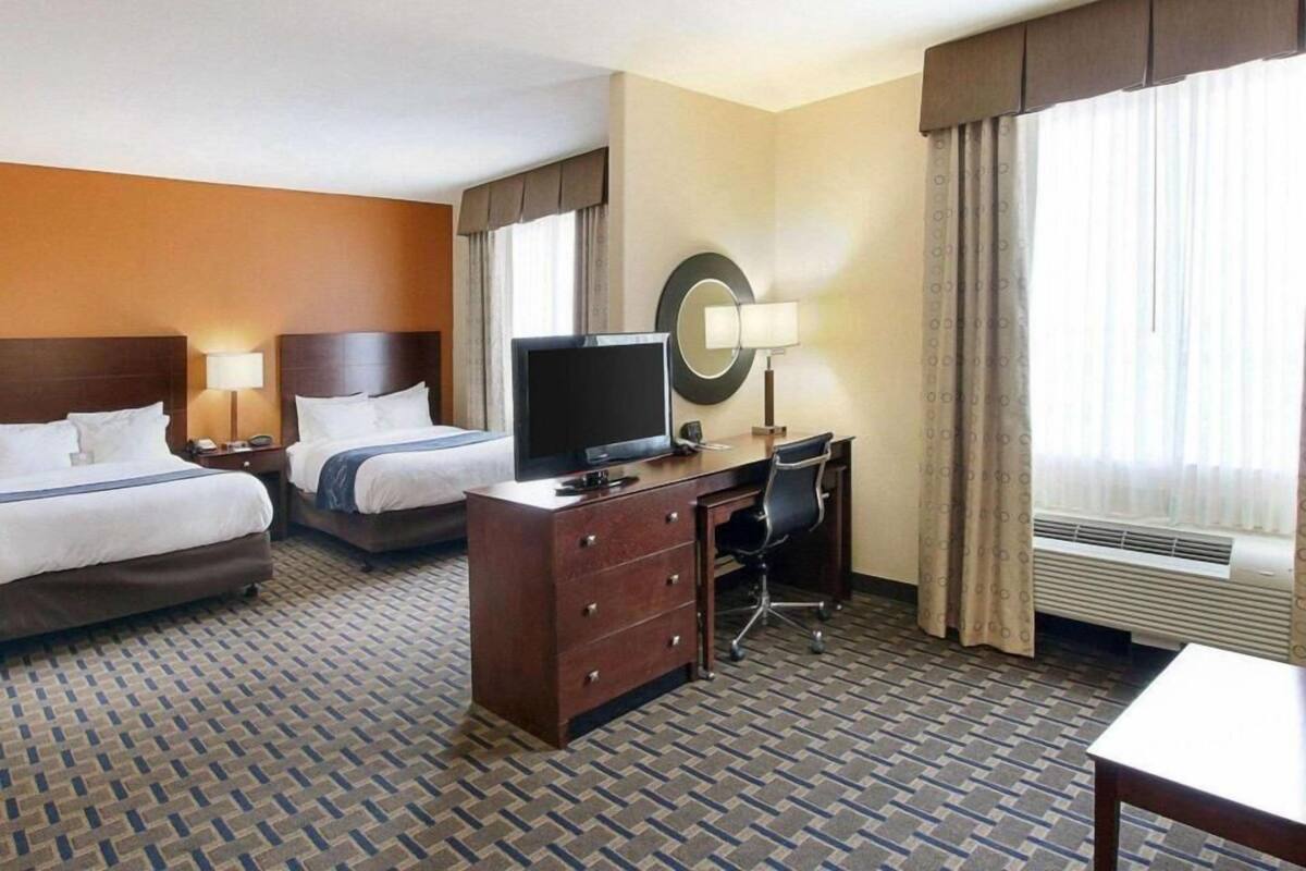Ein Hotelzimmer mit zwei Betten, einem Schreibtisch mit Stuhl, einem Fernseher, einem Spiegel und einem Fenster mit Vorhängen.