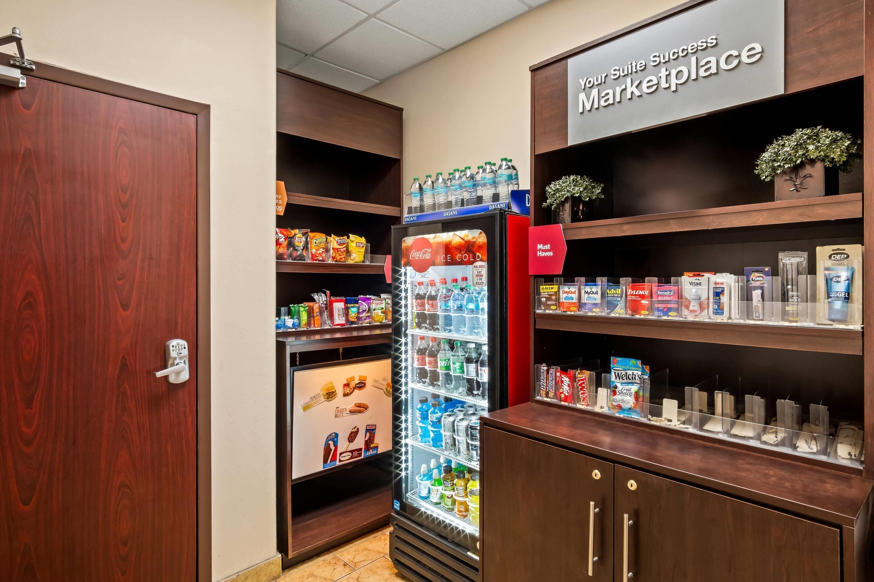 Habitación de hotel con minibar, nevera llena de bebidas y estantes con aperitivos y artículos de aseo.