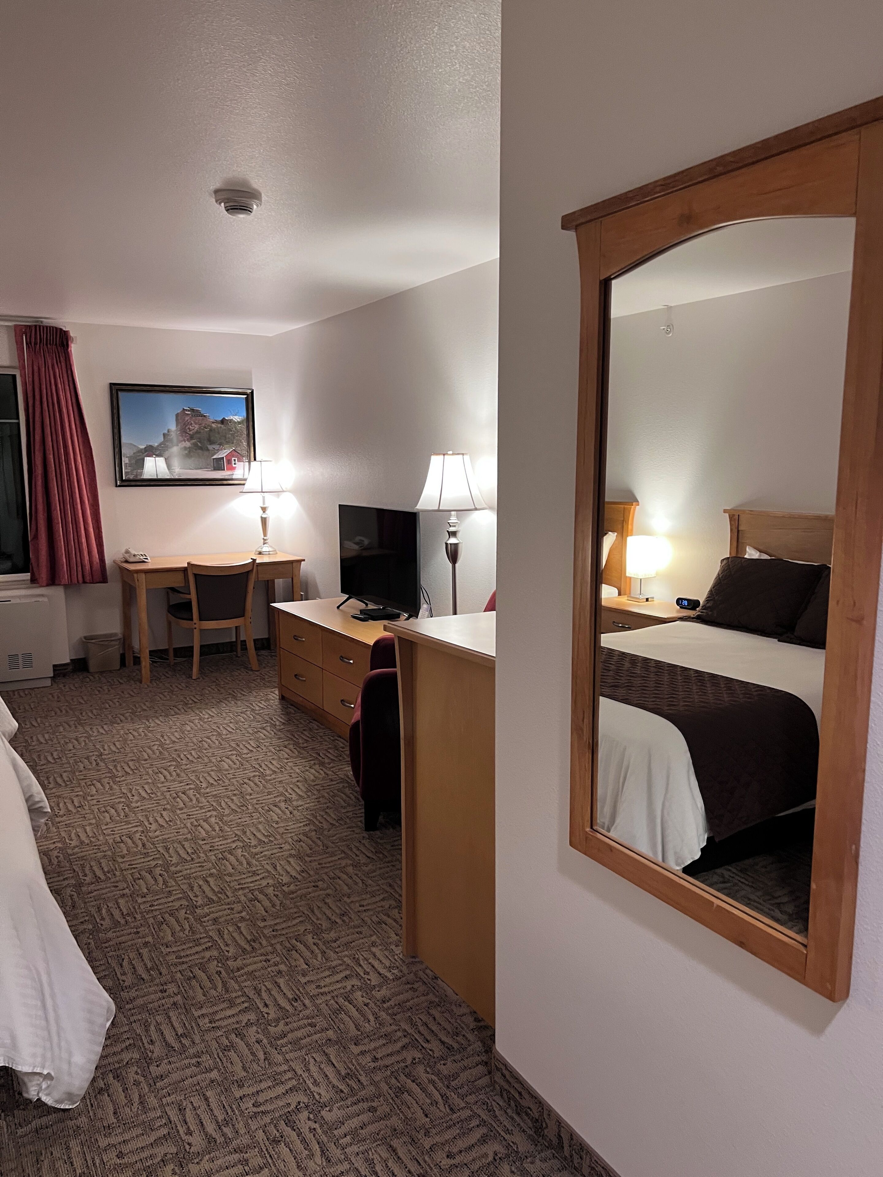 A hotel room with a bed, a desk with a computer, a TV, a lamp, and a large mirror.
