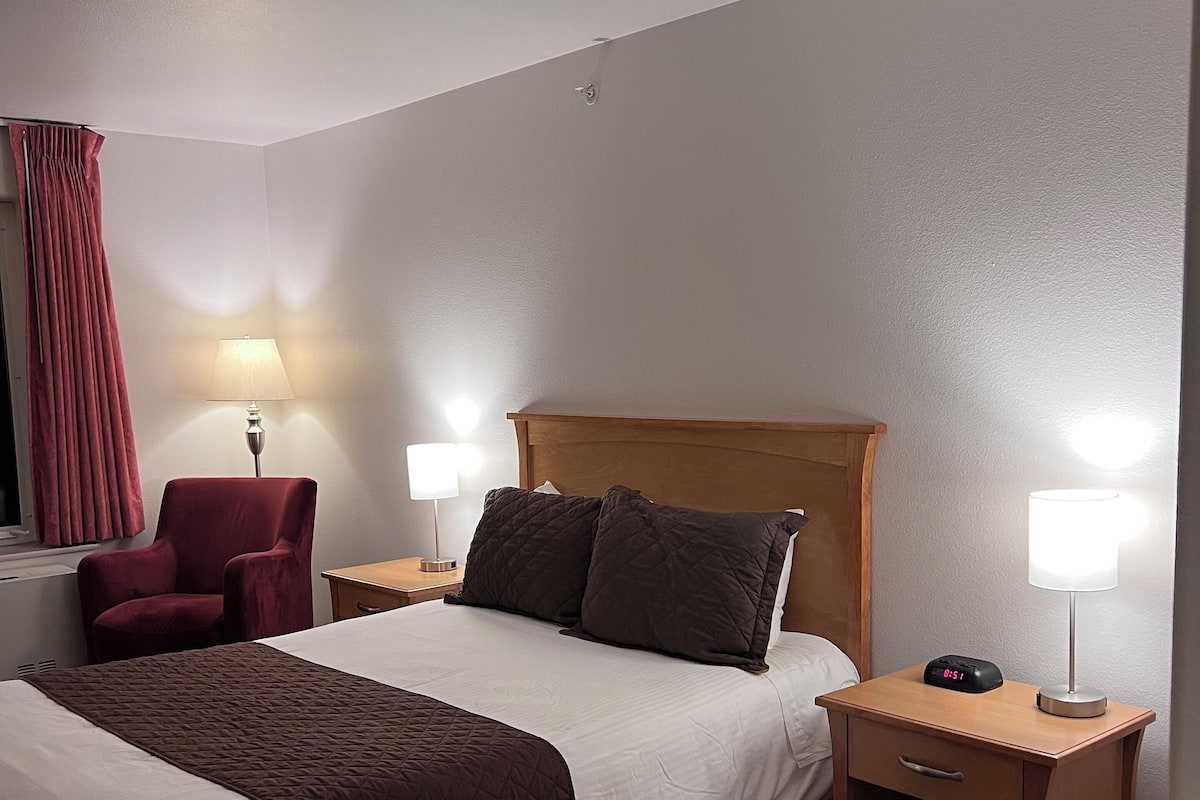 Ein Hotelzimmer mit einem großen Bett, zwei Nachttischen mit Lampen, einem Sessel und einem roten Vorhang.