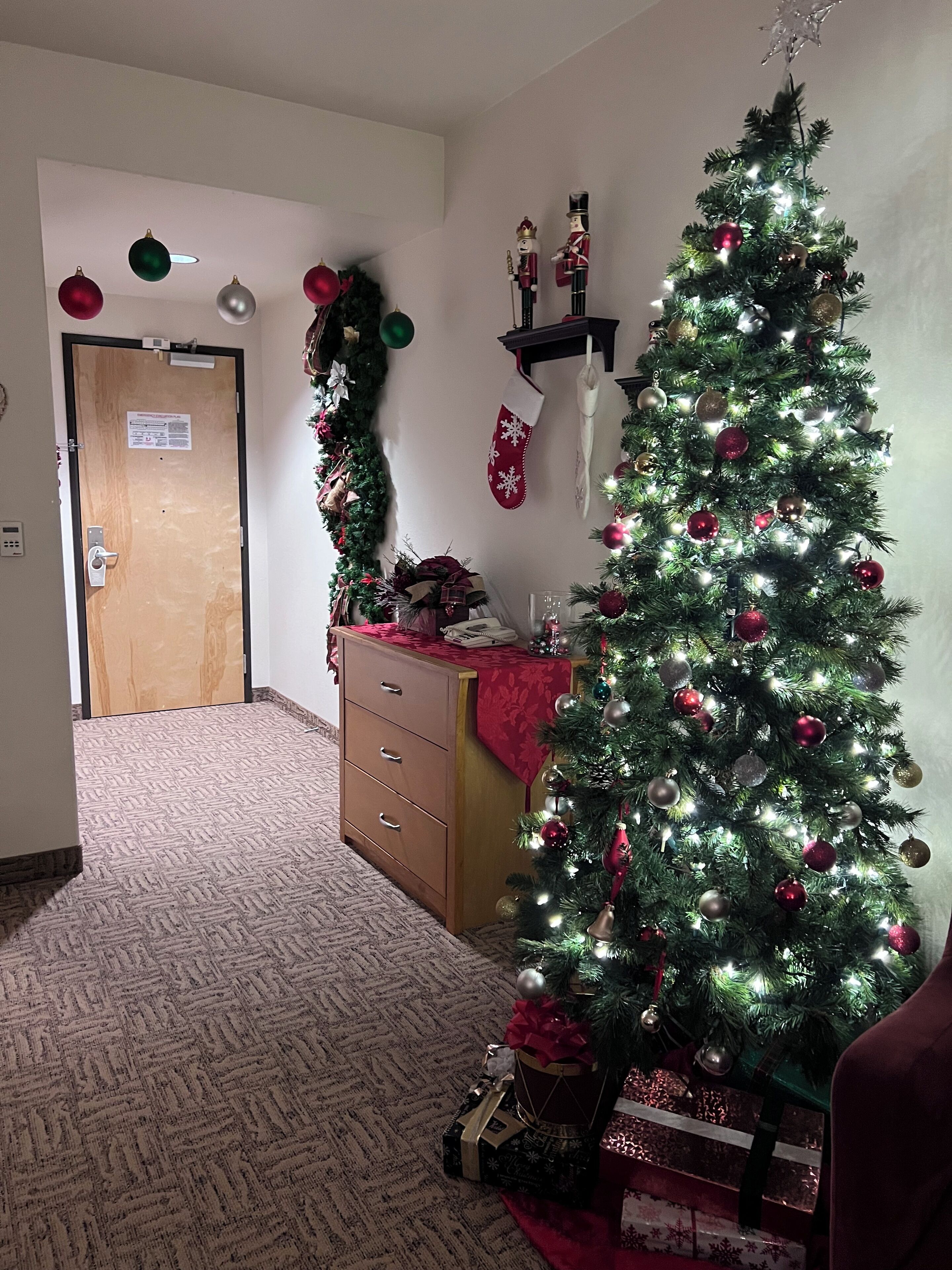 Ein Zimmer mit Weihnachtsbaum, einer Tür, einem Schmuckschrank und einer Wand mit Girlande und Weihnachtssocken.