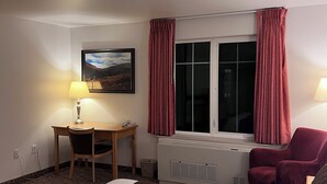 Et hotelværelse med seng, skrivebord, stol, et vindue med gardiner, en radiator og et billede på væggen.