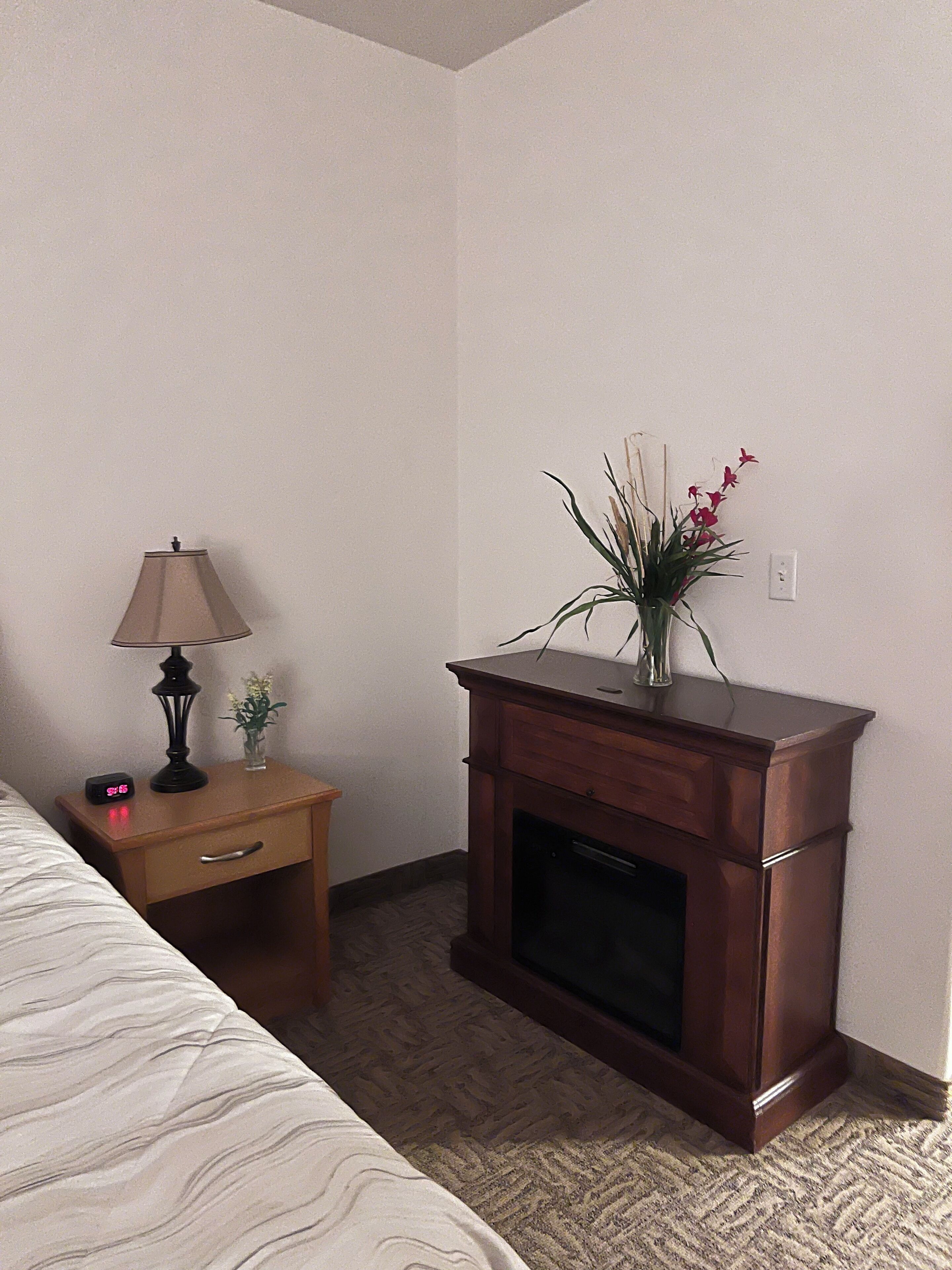 Habitación de hotel con cama, mesita de noche con lámpara, una pequeña mesa con un florero y chimenea.