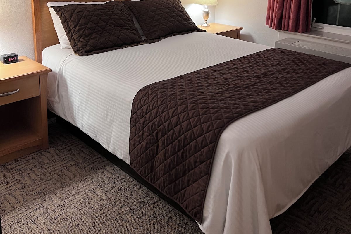 Ein Hotelzimmer mit Bett, Nachttisch, Lampe, Fenster mit Vorhängen und Wanduhr.