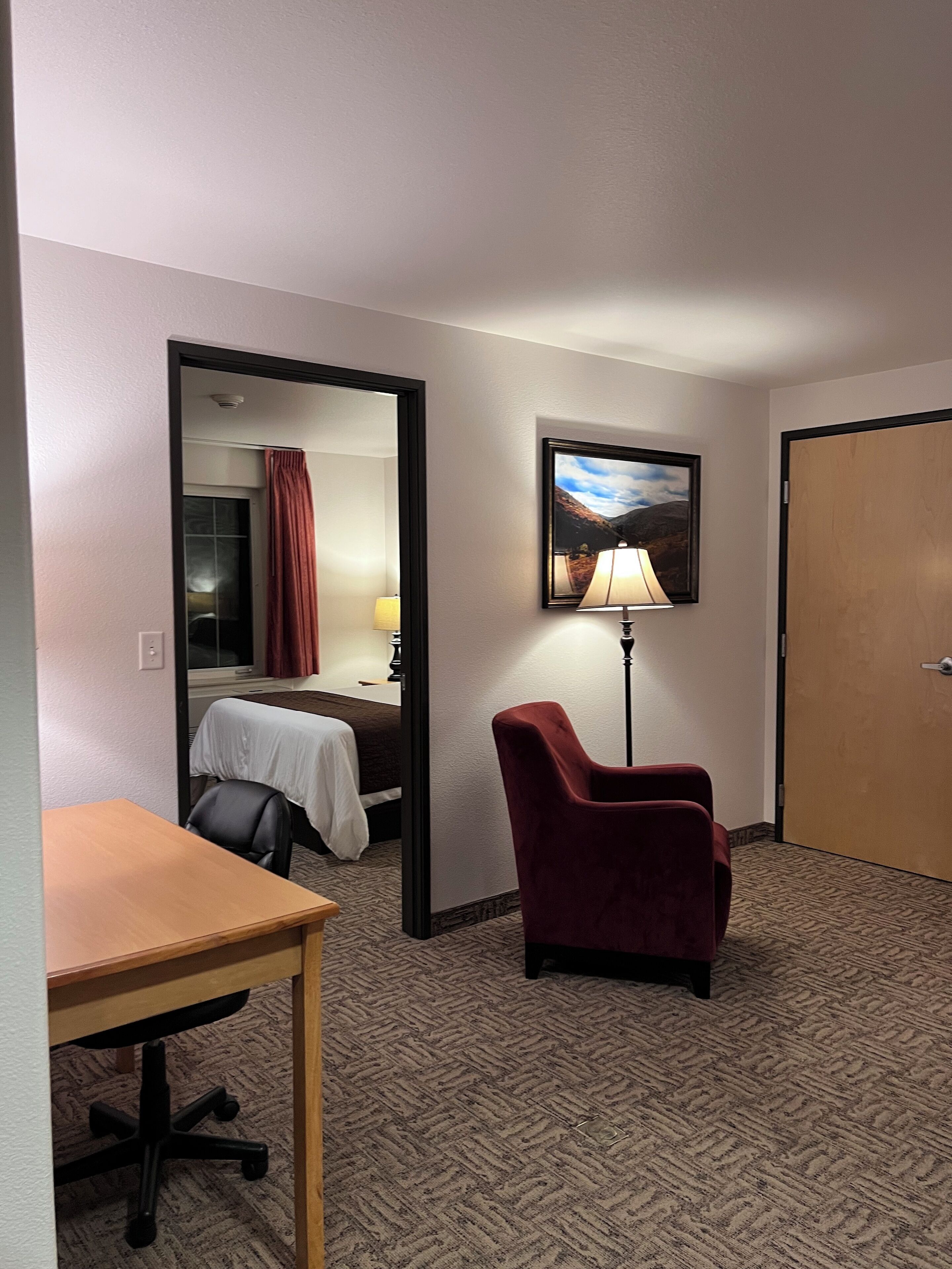 A hotel room with a bed, a desk, a chair, a lamp, and a door.