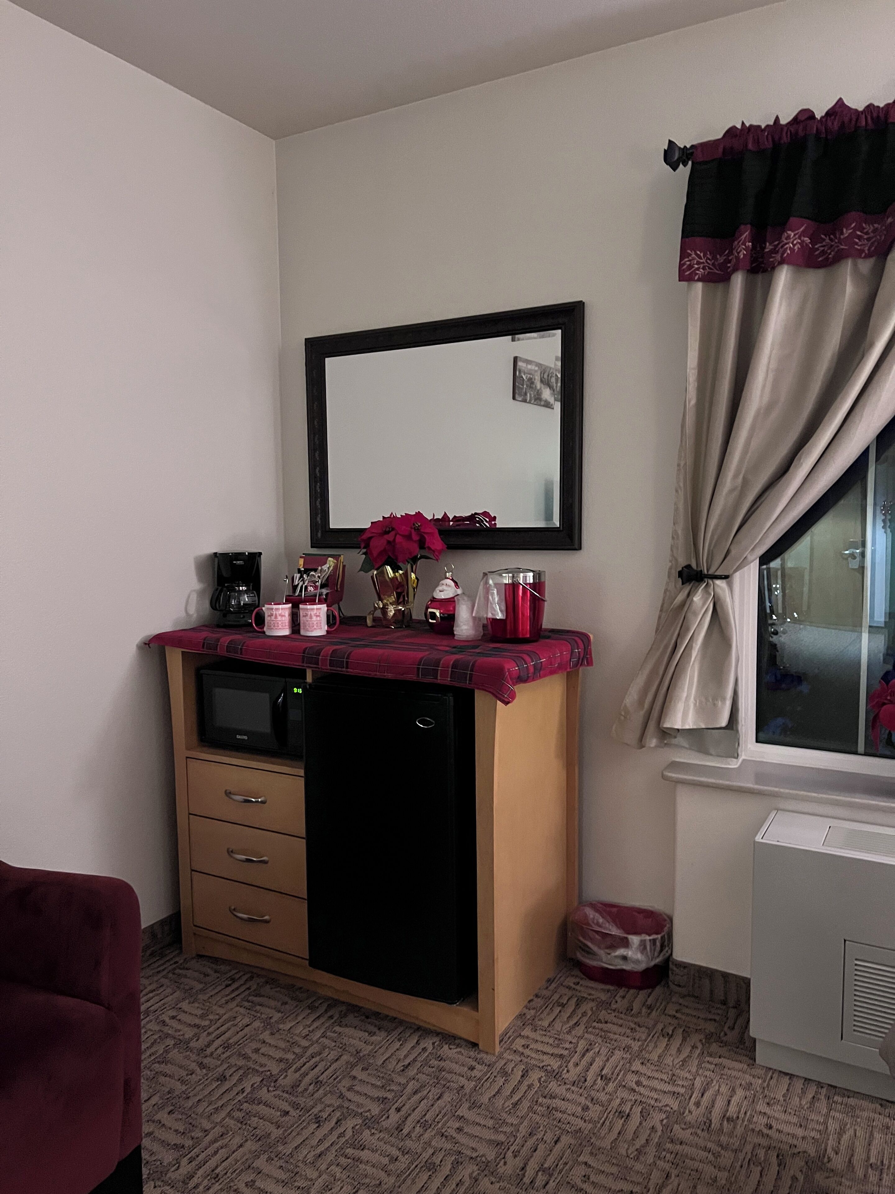 A compact hotel room with a kitchenette, a small refrigerator, a microwave, a coffee maker, and a window with curtains.