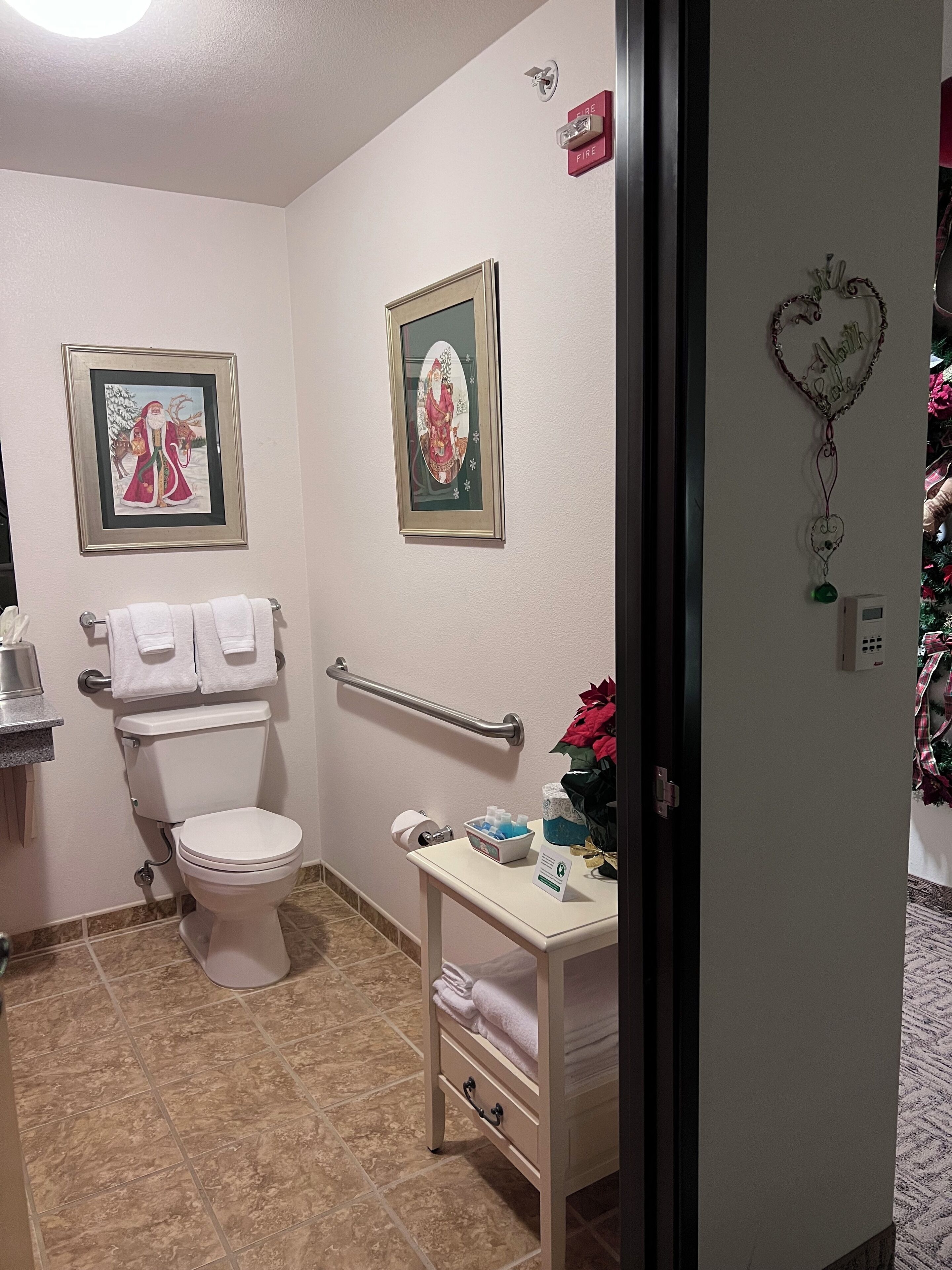 A bathroom with a toilet, a handrail, a framed picture, a small table with towels, and a wall-mounted fire alarm.