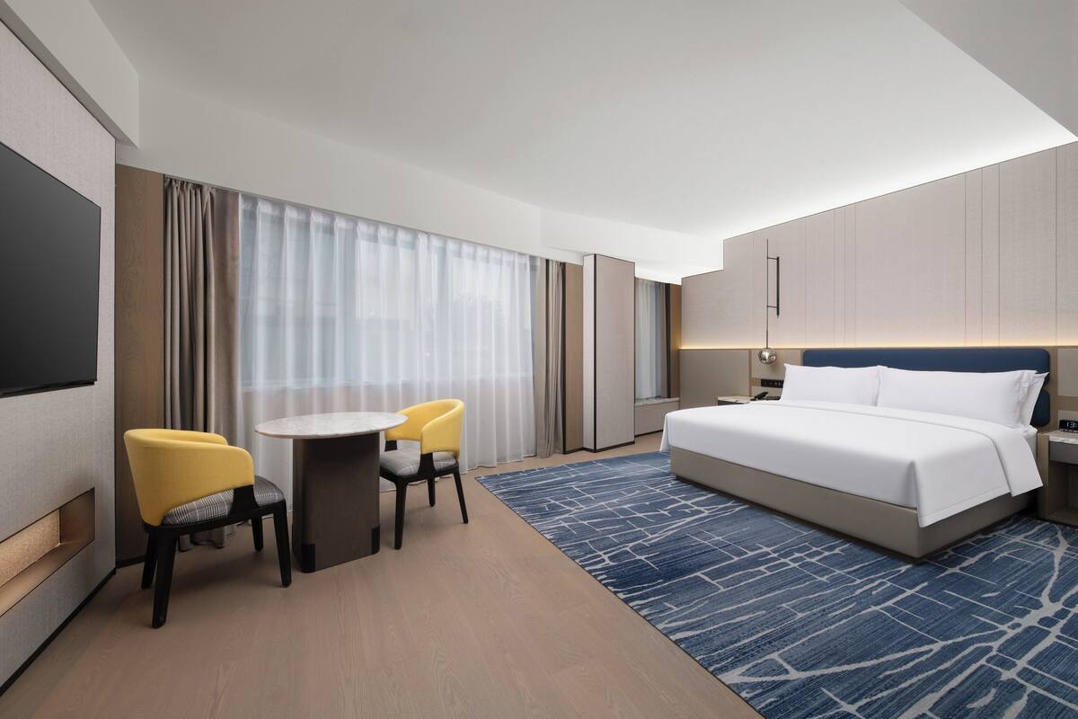 A hotel room with a bed, a desk with two chairs, a TV, and a large window with curtains.