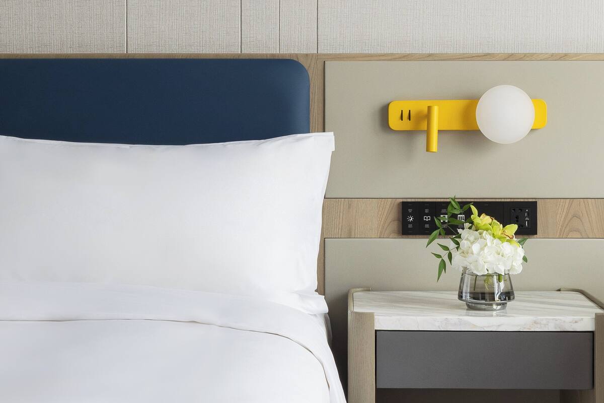 A hotel room with a bed, a nightstand with a vase of flowers, and a wall-mounted light fixture.