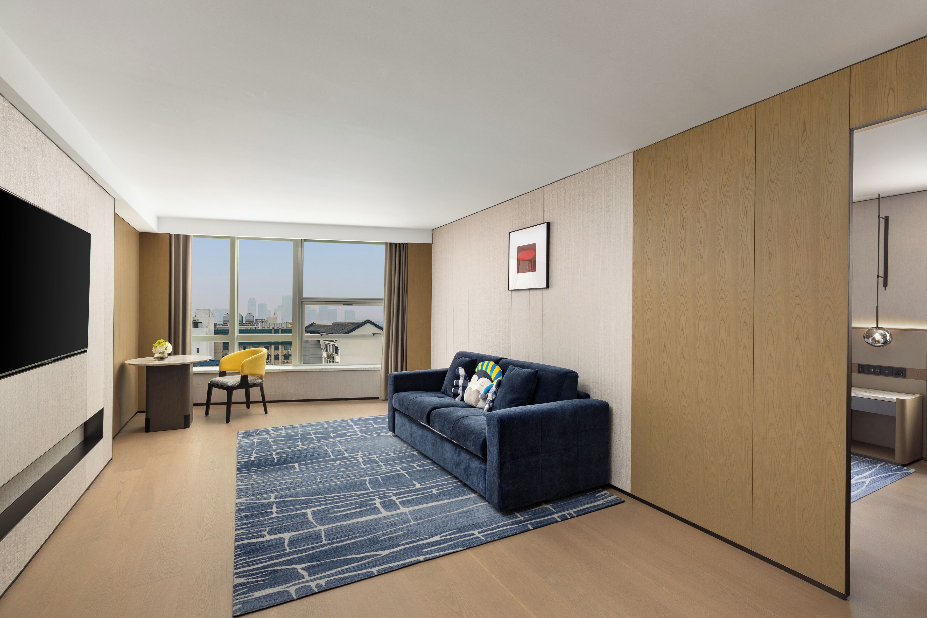 A modern hotel room with a sofa, a small table with a vase, a chair, and a large window offering a city view.