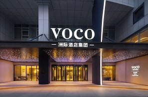 Ein moderner Hotel Eingang mit einem gut sichtbaren 'VOCO'-Schild, Glastüren und einer gut beleuchteten Fassade.
