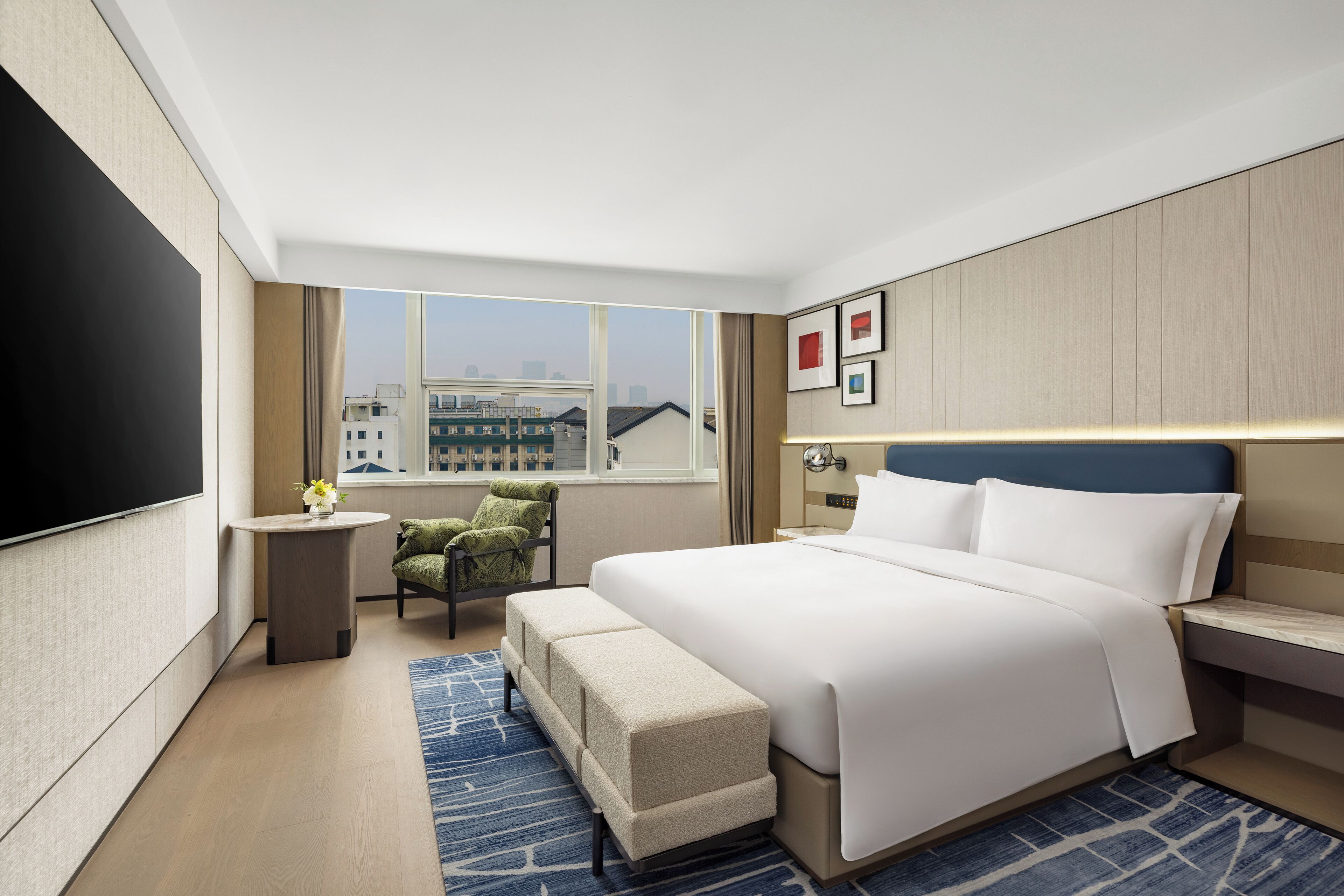 Une chambre d’hôtel moderne avec un grand lit, un téléviseur à écran plat, une petite table ornée d’un vase de fleurs, une chaise et une vue sur la ville par la fenêtre.