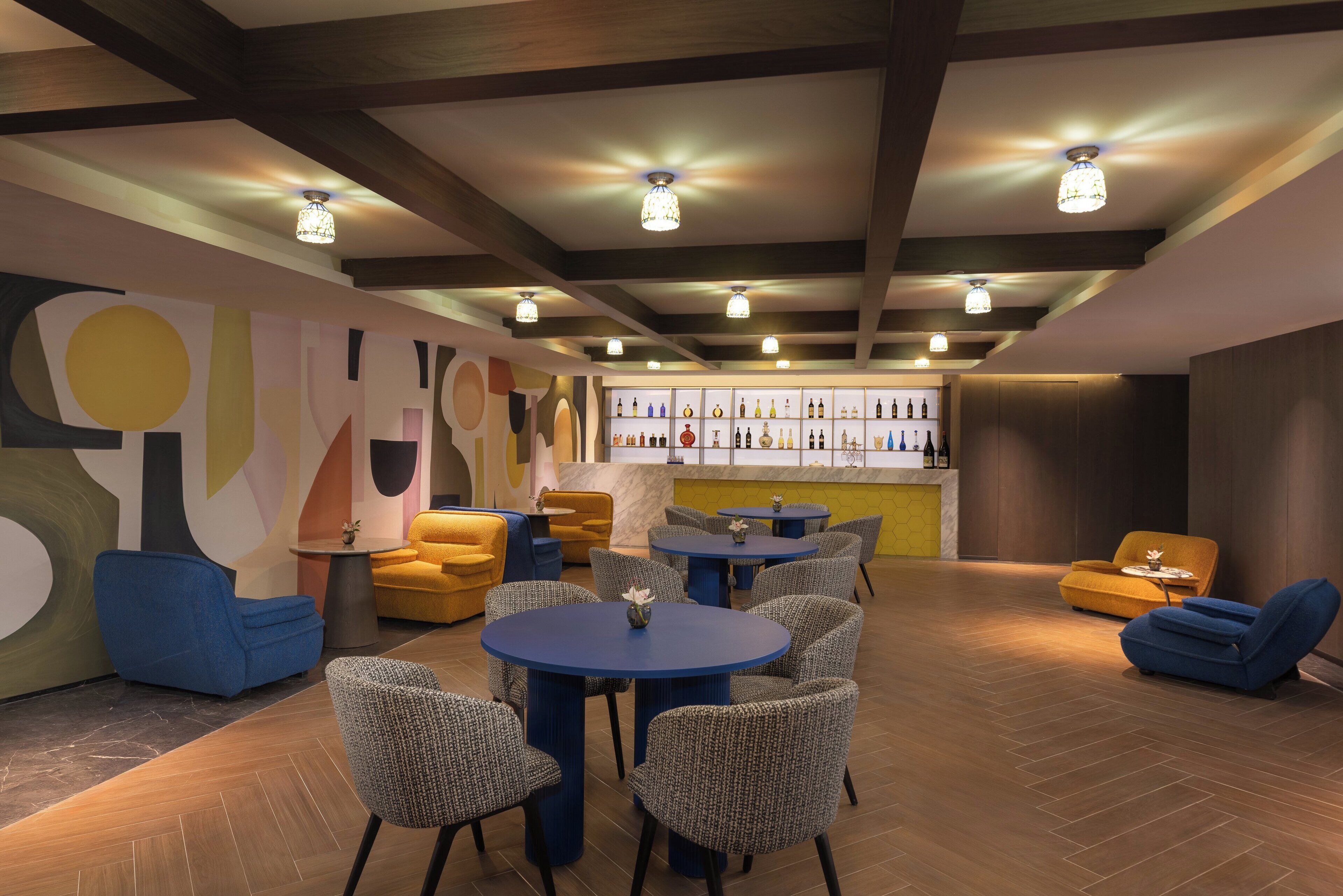A modern lounge area with round tables and comfortable seating, a bar with a variety of bottles, and a colorful abstract wall.
