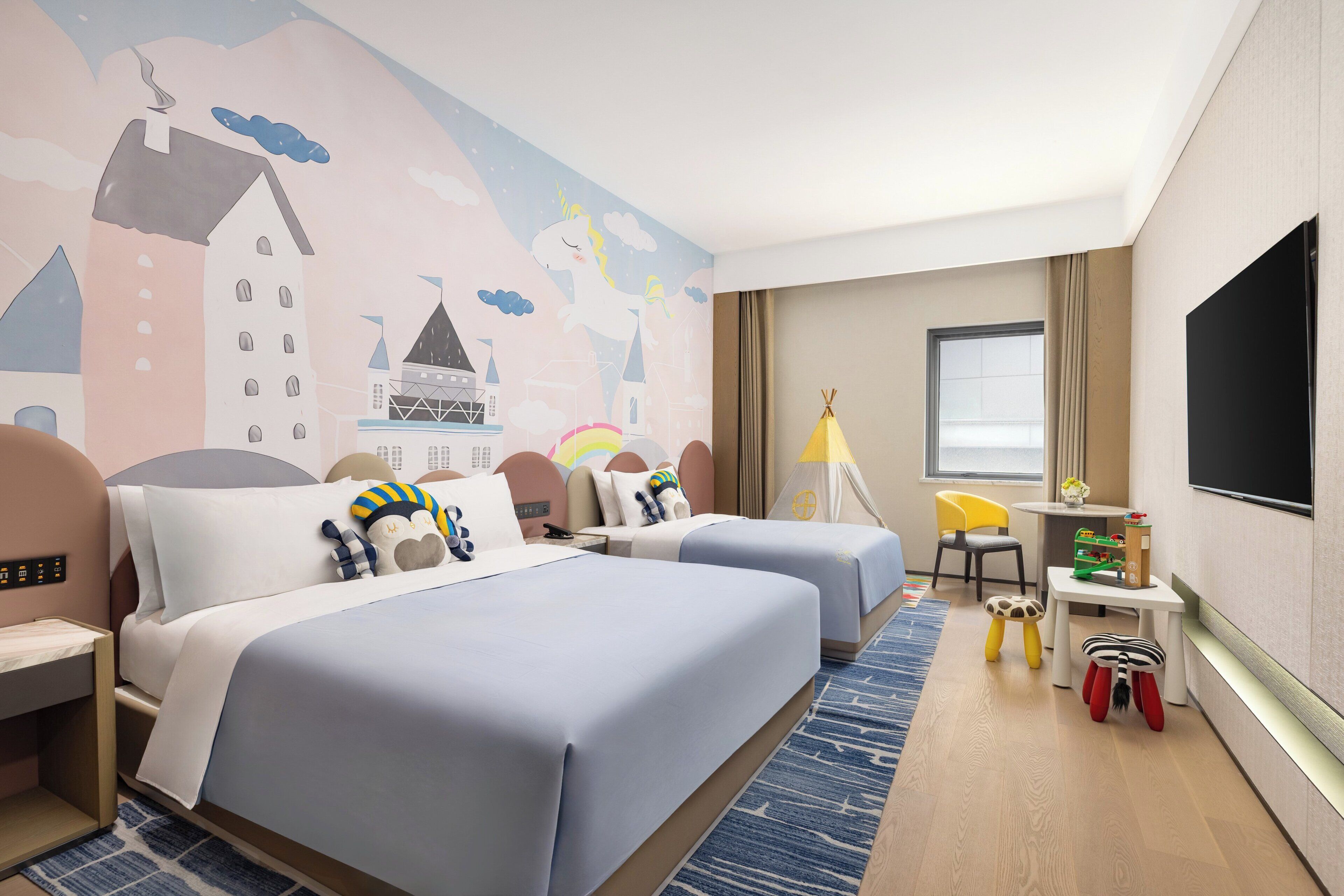 A hotel room with two beds, a TV, a desk, a chair, and a mural on the wall.