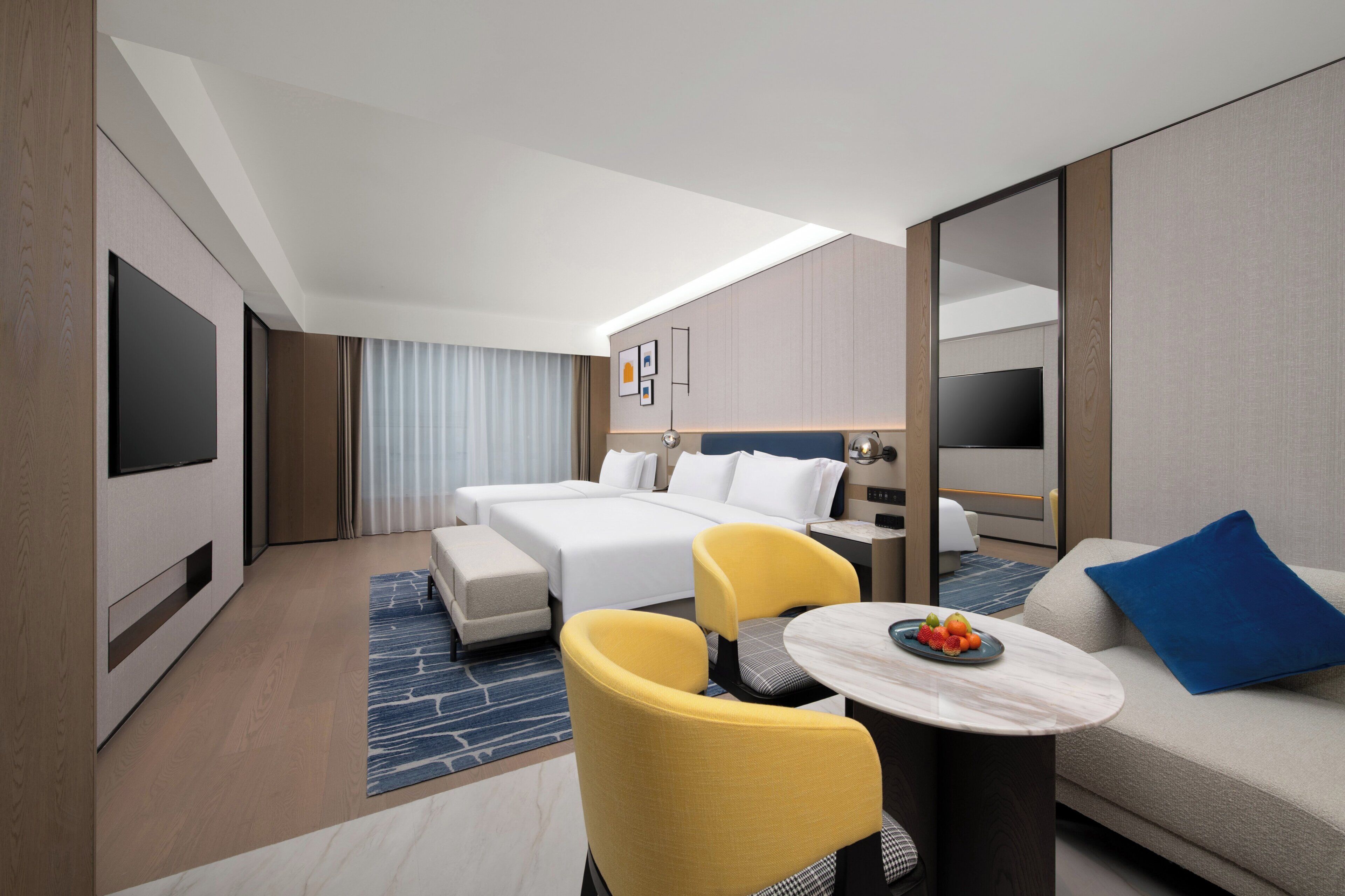 Una habitación de hotel moderna con cama, sofá, una mesita con fruta, una silla y televisor.