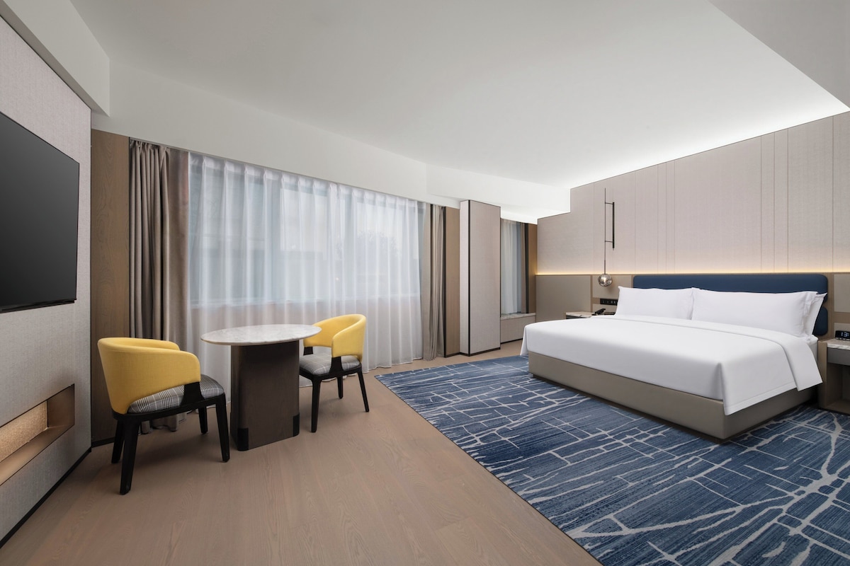 A hotel room with a bed, a desk with two chairs, a TV, and a large window with curtains.