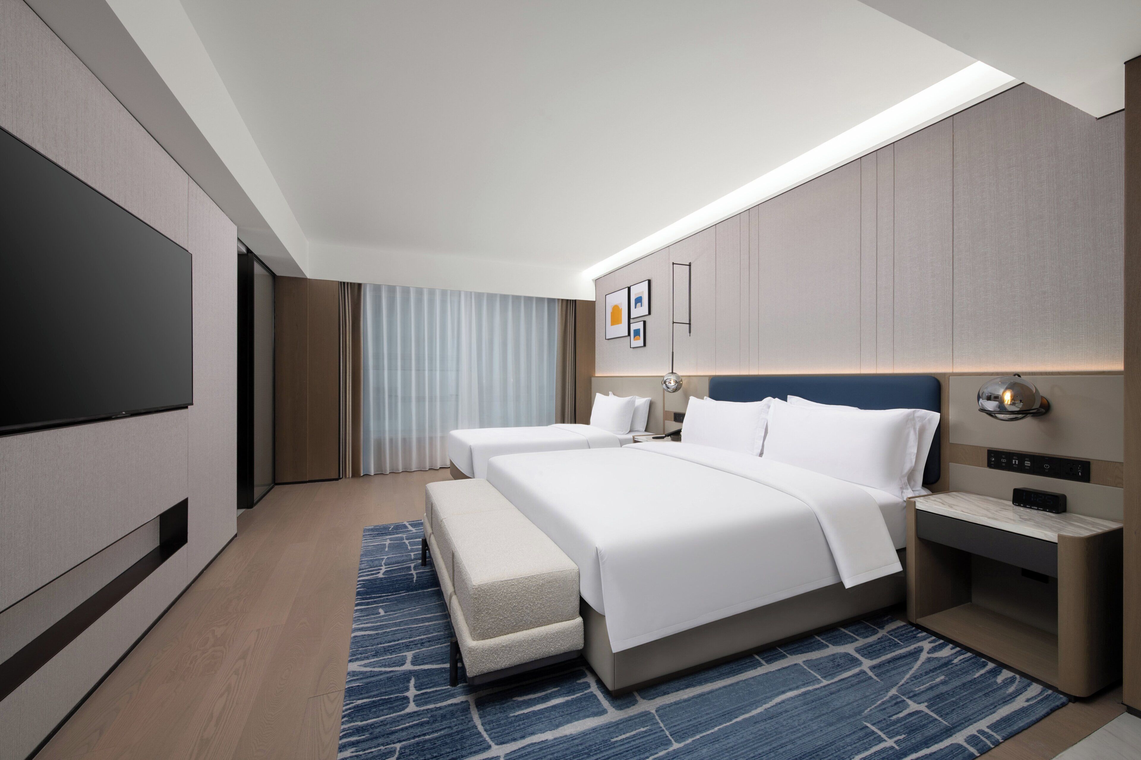 A modern hotel room with a large bed, a flat-screen TV, a bedside table, and a desk with a lamp.