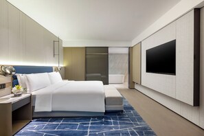 A modern hotel room with a large bed, a flat-screen TV mounted on the wall, a bedside table with a lamp, and a bathroom visible through a sliding door. - voco Suzhou Guanqian Street by IHG (Suzhou)