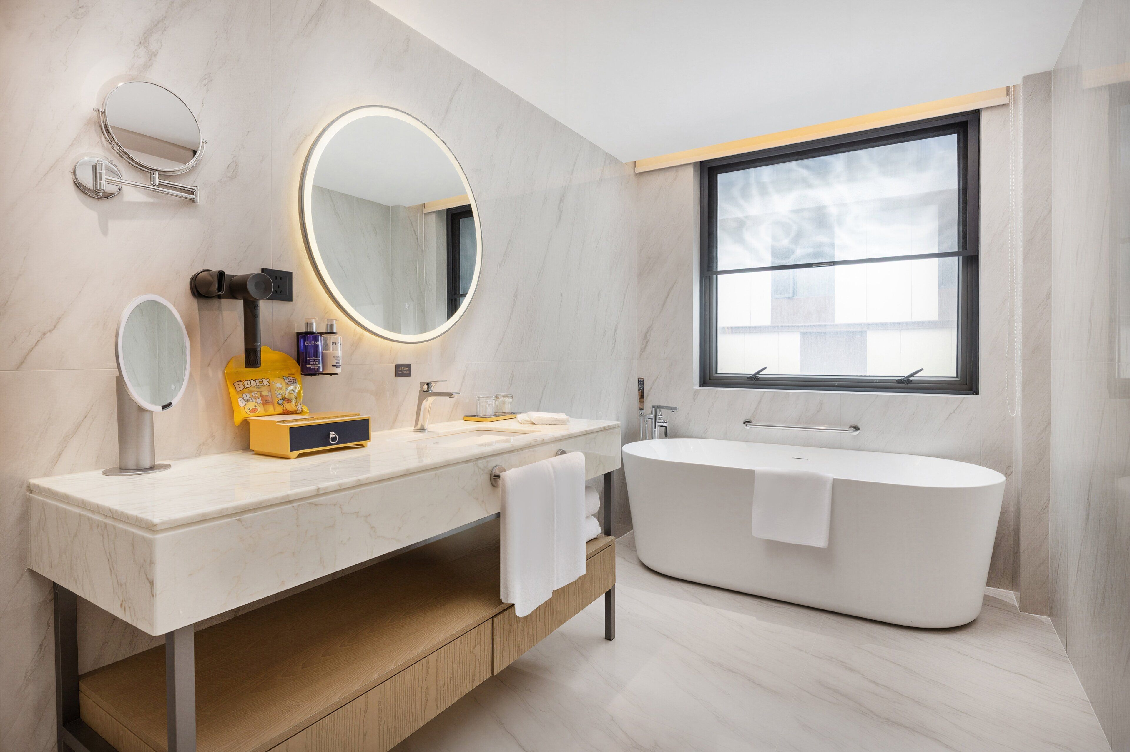 Een moderne badkamer met een vrijstaand bad, een grote spiegel en een raam dat voor natuurlijk licht zorgt.