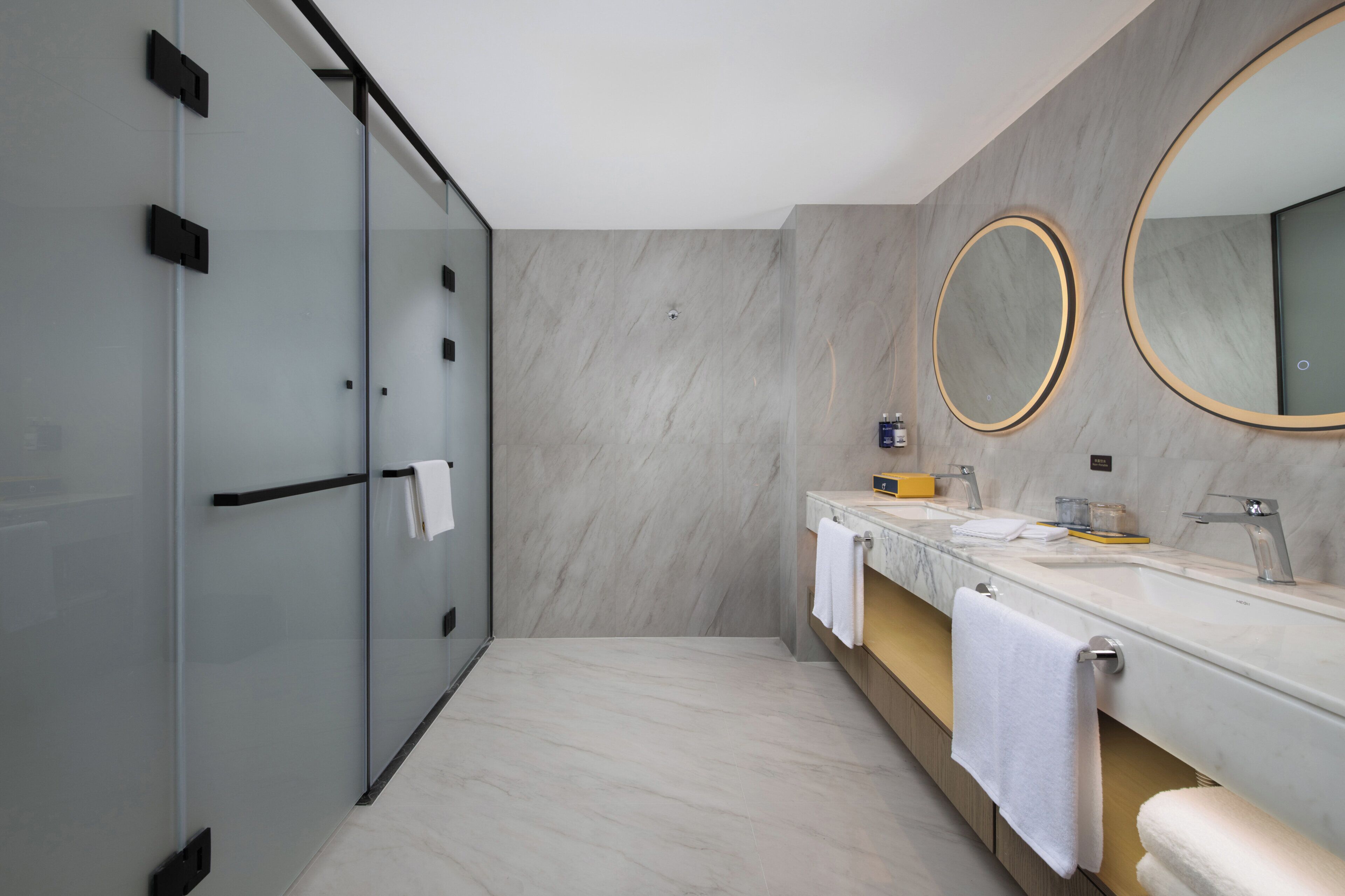 Un baño moderno con cabina de ducha de vidrio, tocador de mármol con dos lavamanos y dos espejos grandes.