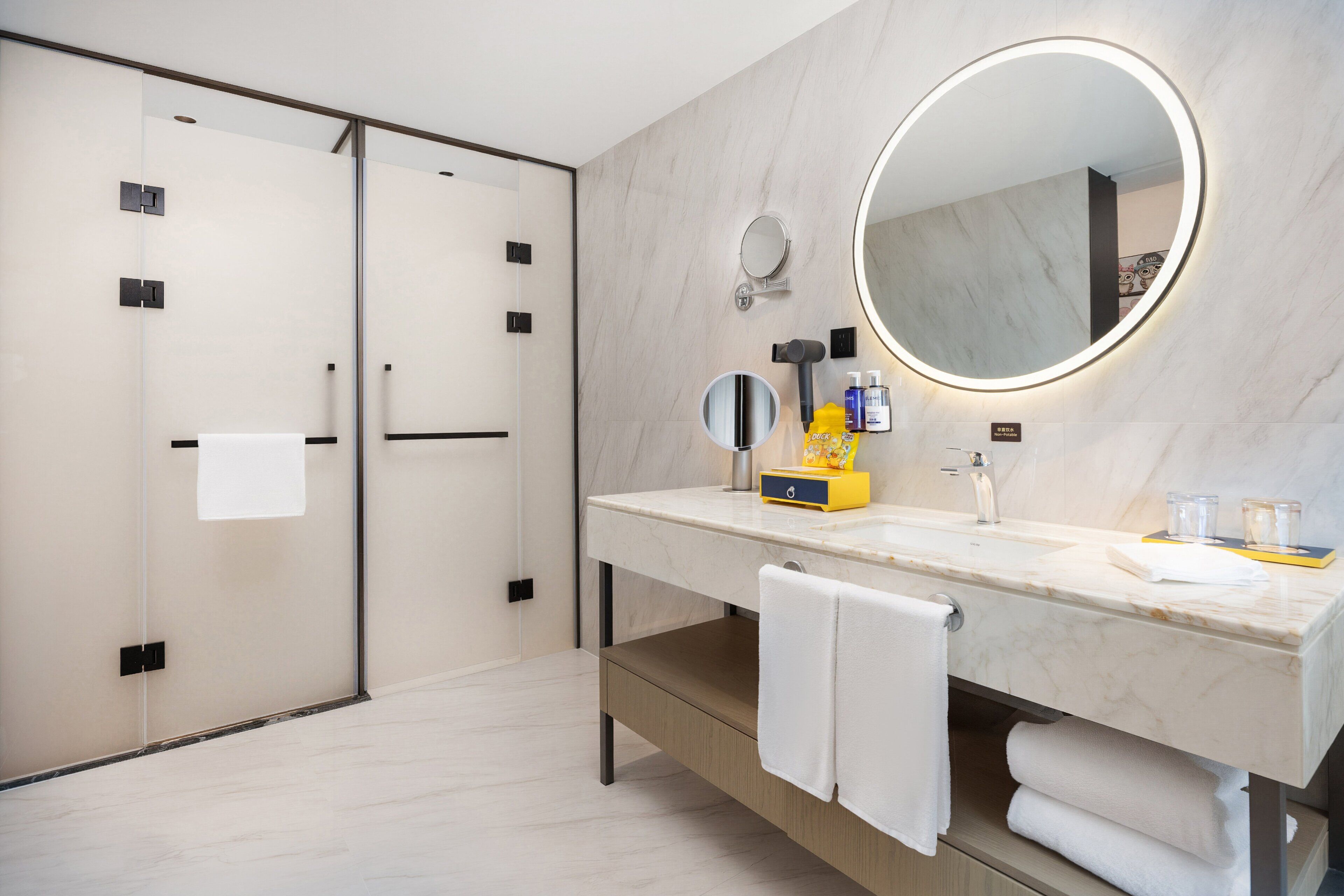 A modern bathroom with a marble countertop, a round mirror with a light, a towel rack, and a glass shower enclosure.