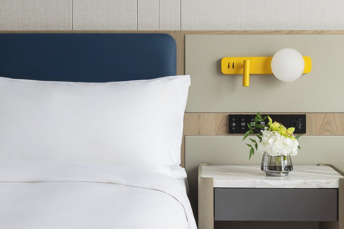 A hotel room with a bed, a nightstand with a vase of flowers, and a wall-mounted light fixture.