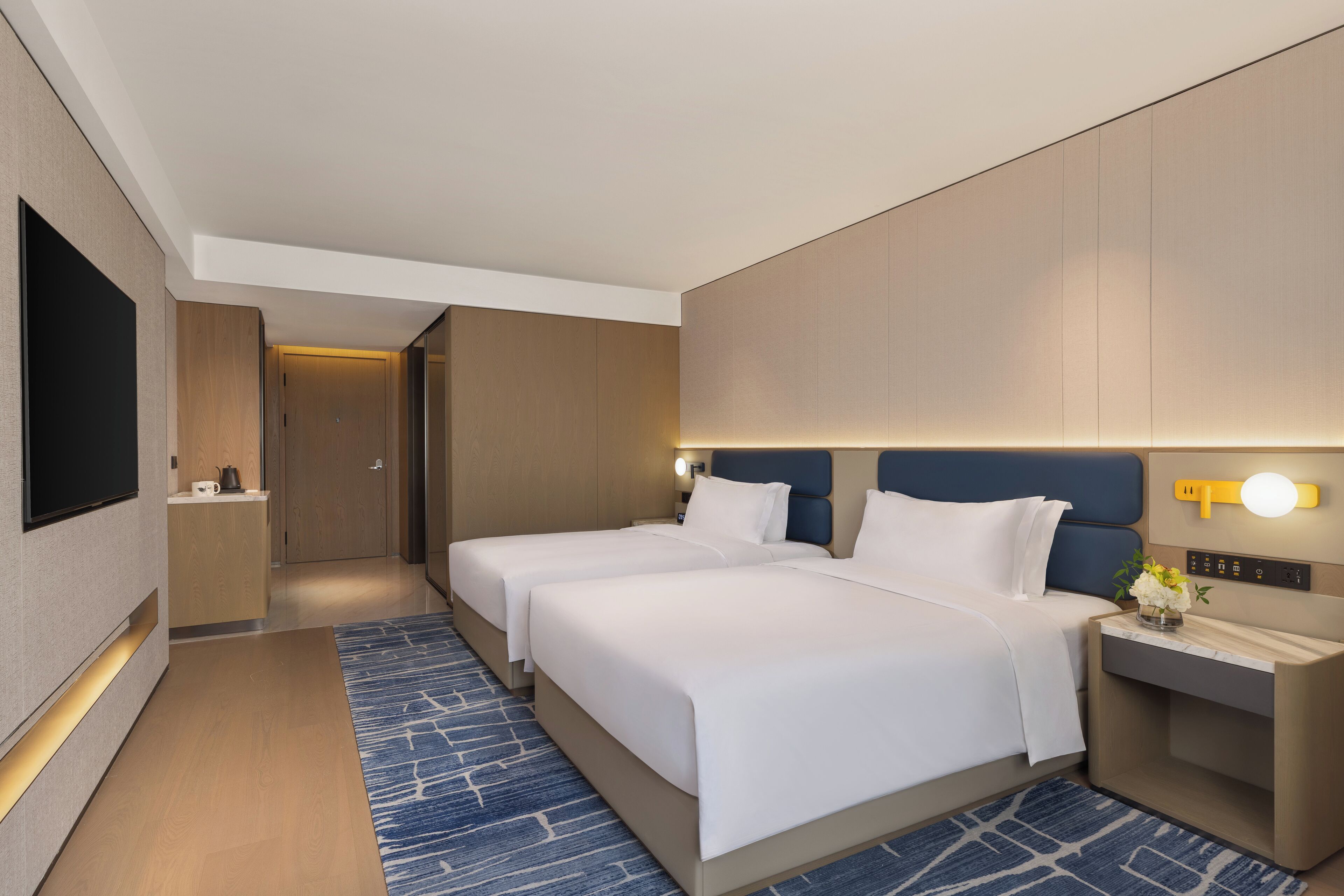 A hotel room with two beds, a TV, a bedside table with a lamp, and a small desk with a flower vase.