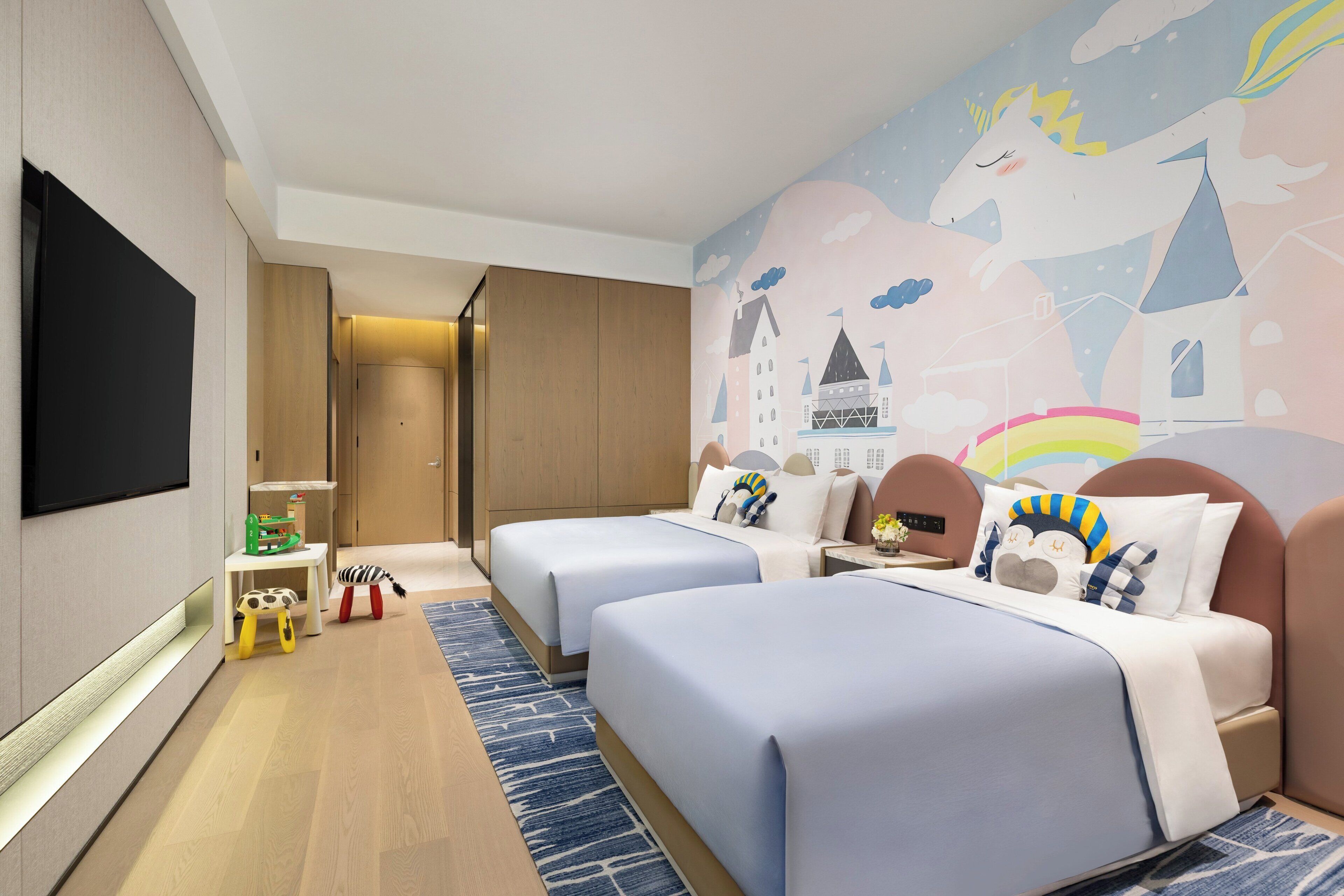 Une chambre d’hôtel avec deux lits, un téléviseur à écran plat, une armoire en bois, un bureau avec une lampe et une fresque colorée sur le mur.