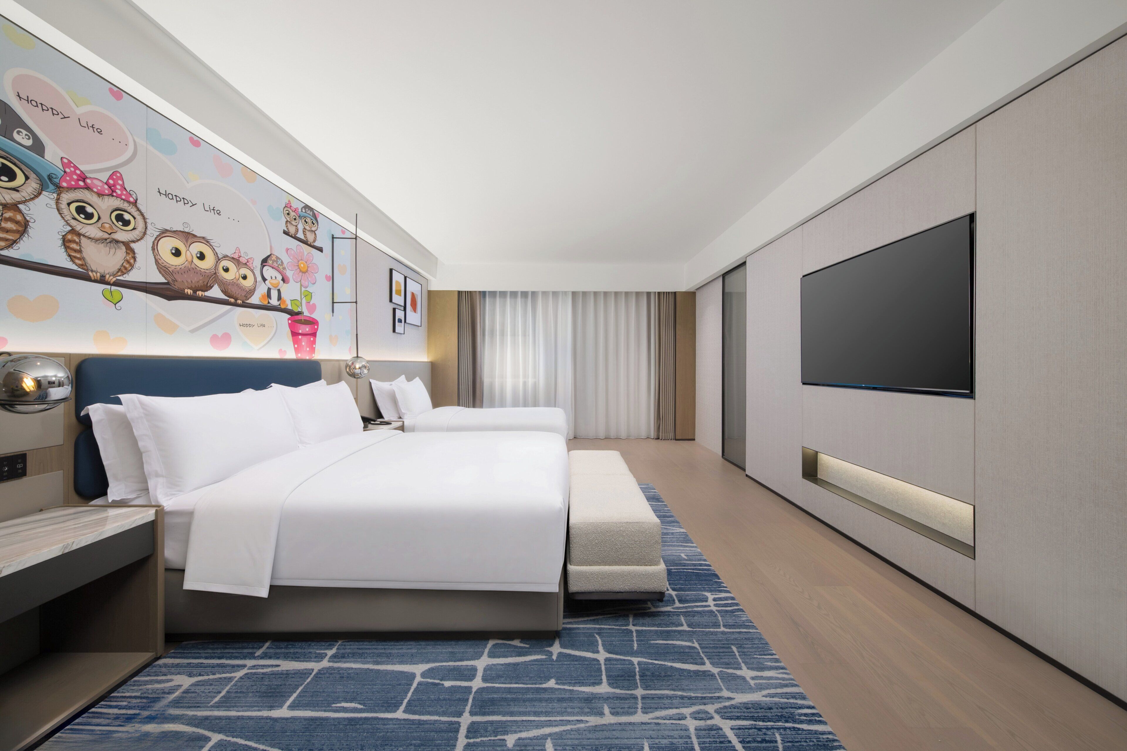 A modern hotel room with a large bed, a flat-screen TV, a desk, and a wall with owl illustrations.
