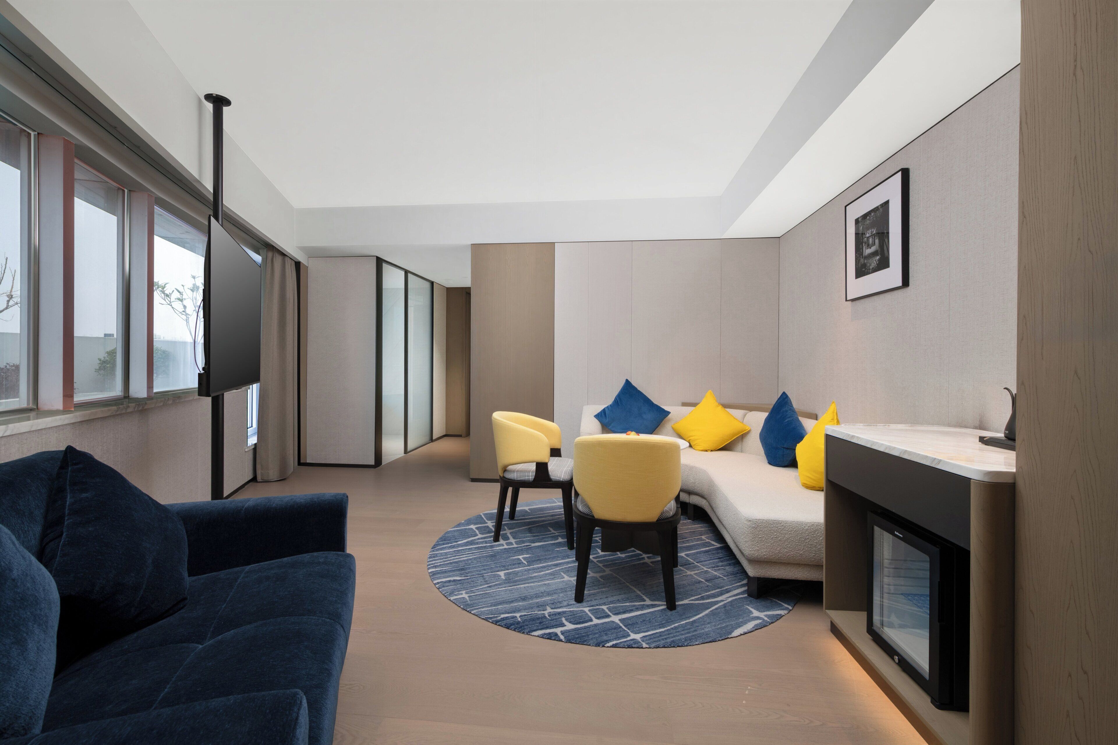 A modern hotel room with a sofa, chairs, a small table, a fireplace, and large windows.