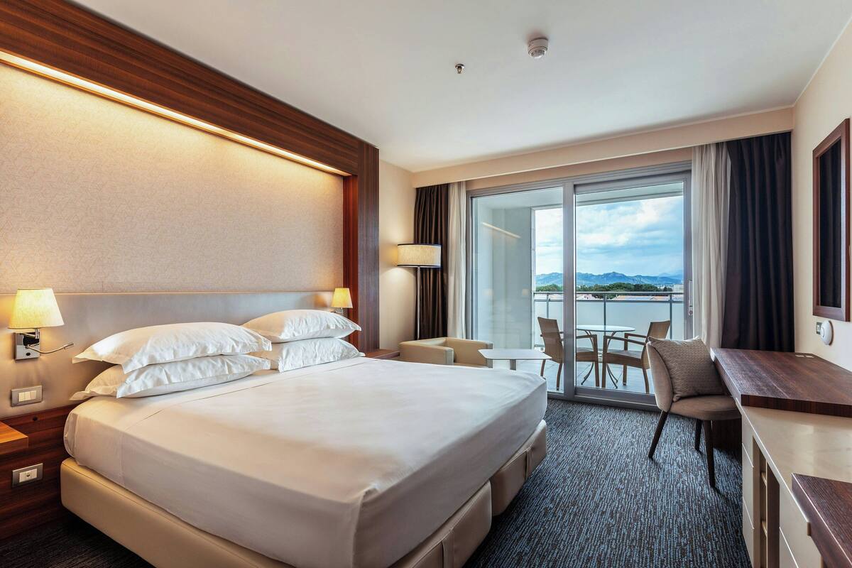 A hotel room with a large bed, a desk, a chair, and a view of the outdoors.