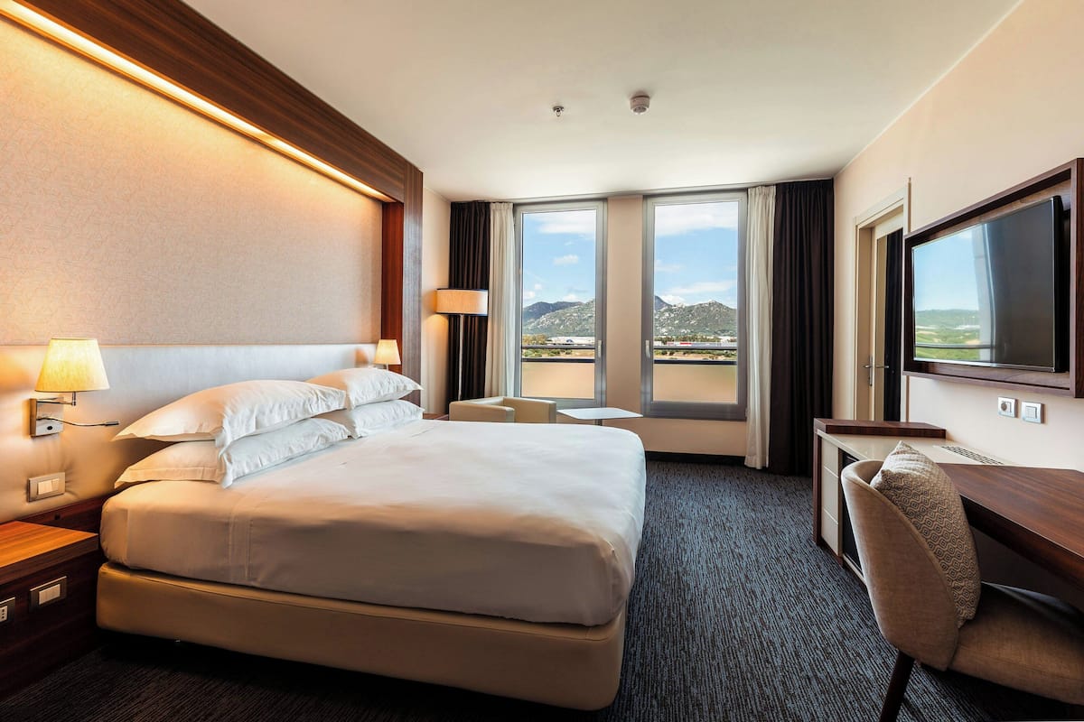 A hotel room with a large bed, a desk, a chair, a TV, and a view of the mountains.