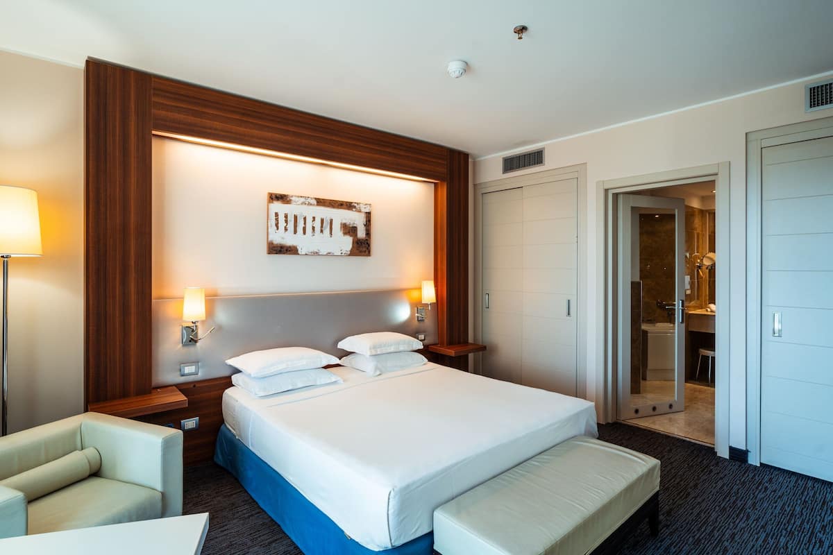 A hotel room with a large bed, bedside lamps, a wooden headboard, a bench, and a door leading to a bathroom.