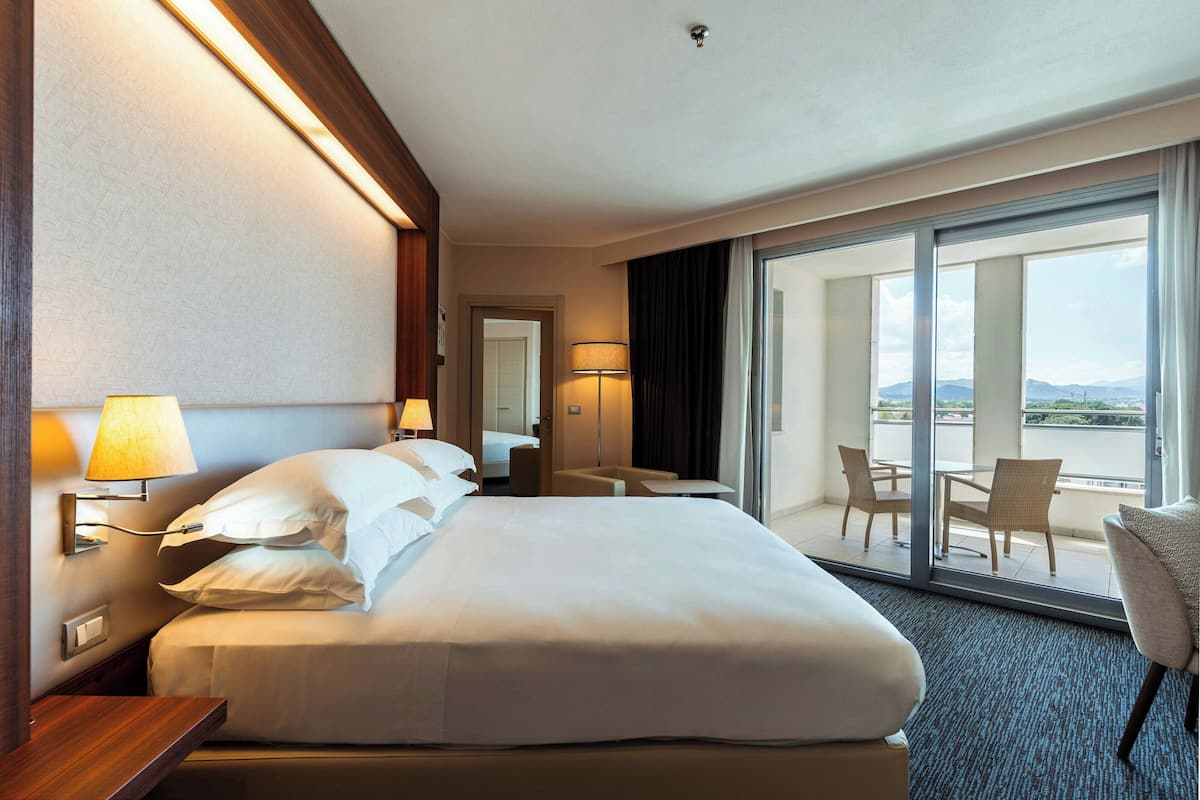 A hotel room with a large bed, a desk, a chair, and a balcony with a table and chairs.